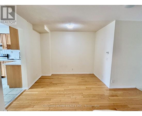 A room with wooden flooring and white walls, with a kitchen area visible in the background.