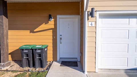 Two green bins are placed on the ground outside a yellow house.