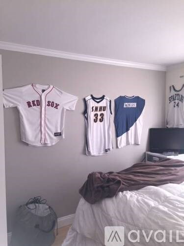 A bedroom with a bed, a wall-mounted jersey rack holding three jerseys, and a backpack on the floor.