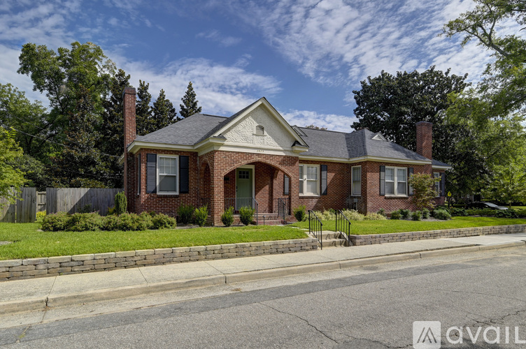 A house with a red brick exterior and a grey roof is surrounded by greenery and a clear sky.