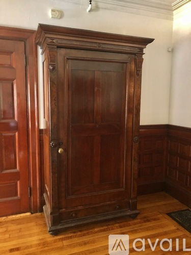 A tall wooden armoire stands in a room with a wooden floor.