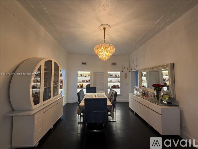 A chandelier hangs from the ceiling in a spacious room with white walls and a long table with chairs.