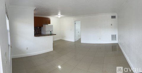 A spacious empty room with white walls and a kitchen area in the background.