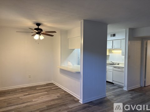A room with a ceiling fan and a kitchen area in the background.