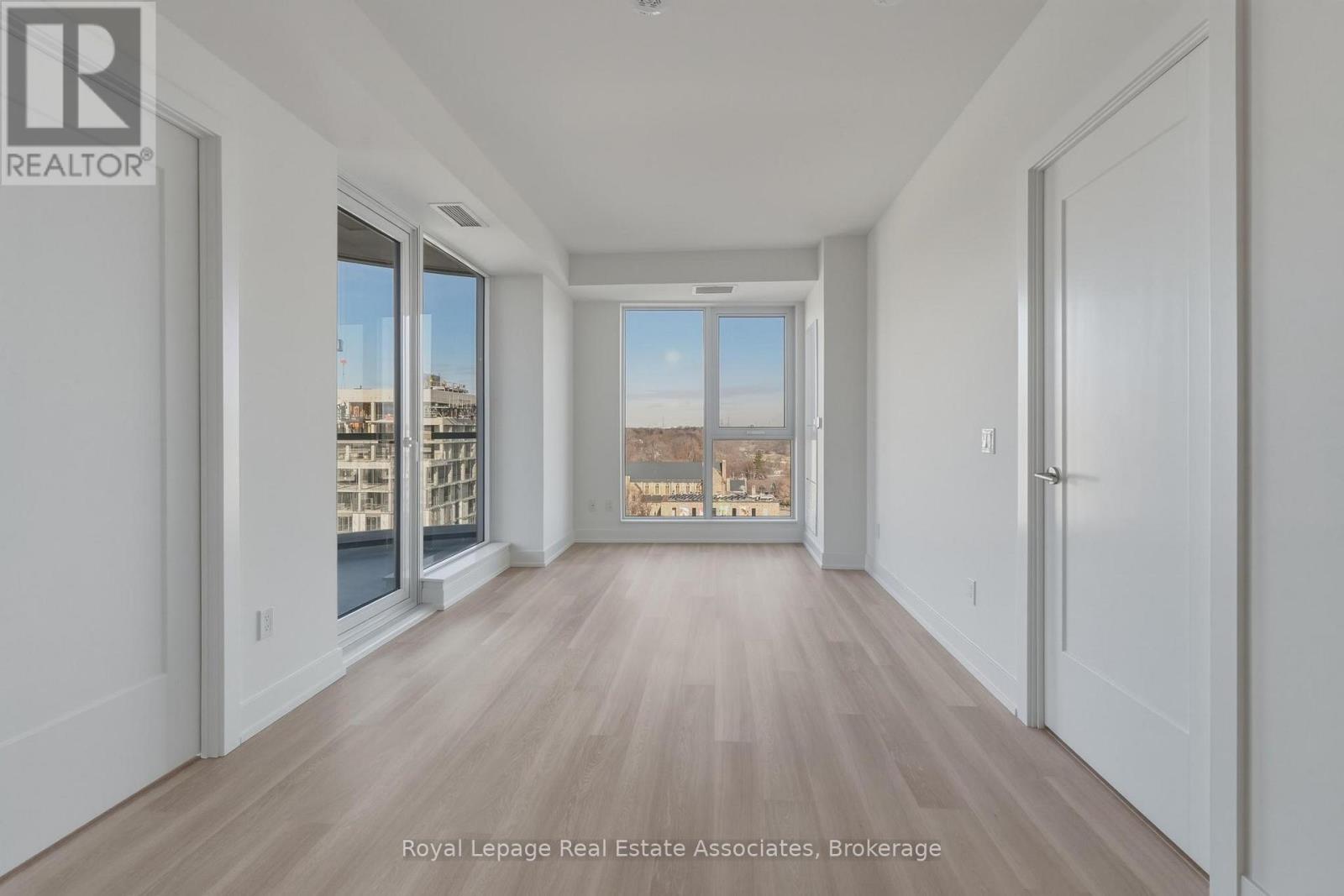 A room with wooden floors and white walls, with a view of buildings outside through the windows.