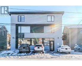 A modern house with a white and black exterior and a large glass window is shown with three cars parked in front.