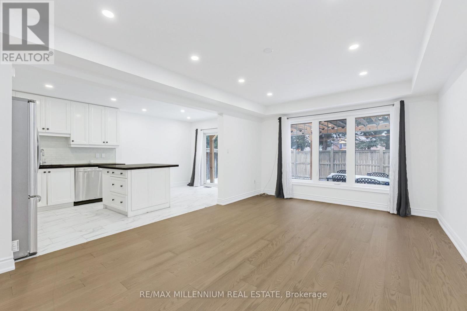 A spacious room with wooden flooring and white walls, featuring a kitchen area with white cabinets and a window with a view of the city.