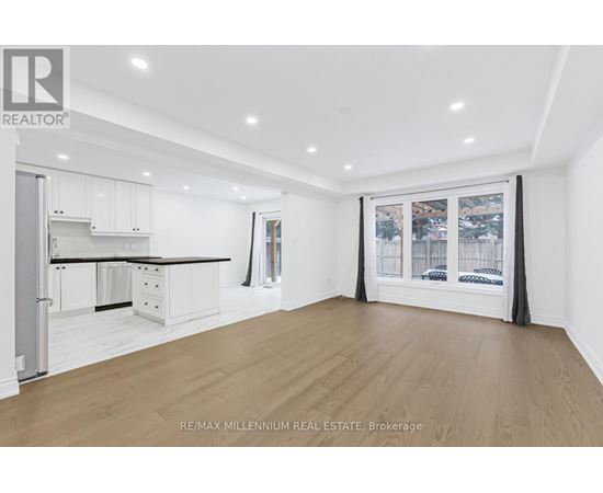 A spacious room with wooden flooring and white walls, featuring a kitchen area with white cabinets and a window with a view of the city.