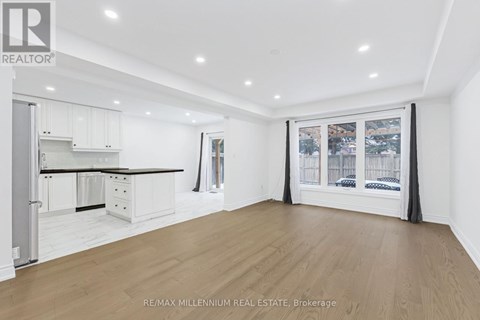 A spacious room with wooden flooring and white walls, featuring a kitchen area with white cabinets and a window with a view of the city.