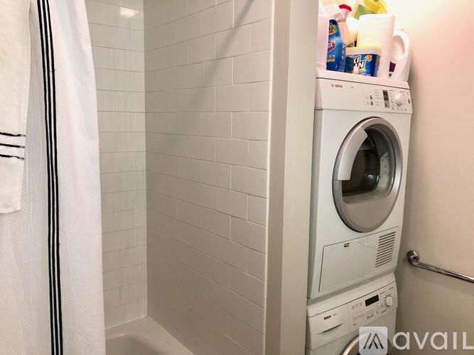 A white washing machine is in a bathroom with a white tiled wall.