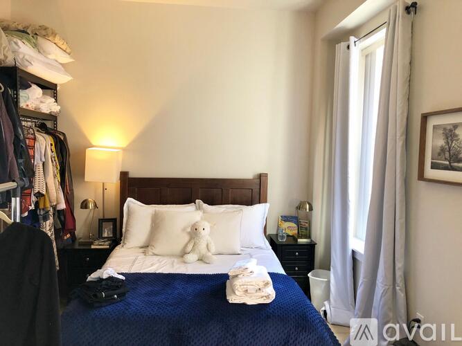 A neatly made bed with a blue comforter and white pillows sits in a bedroom with a window and a clothing rack.