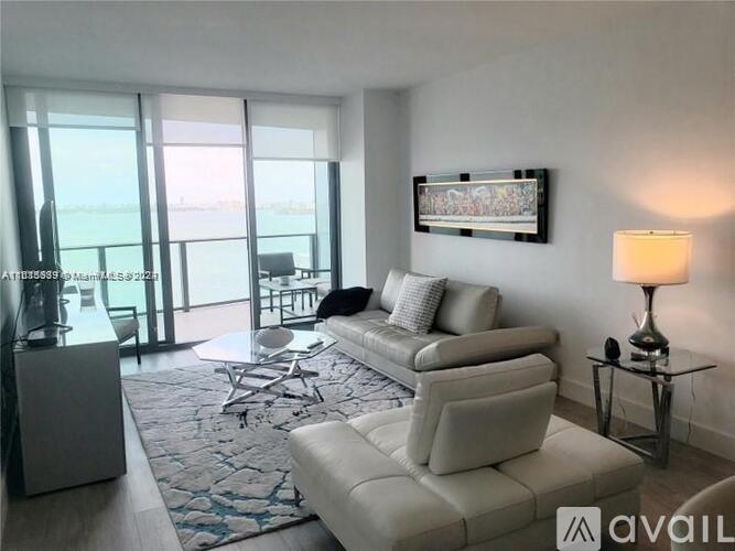 A living room with a white couch, a chair, a table with a lamp, and a flat screen TV mounted on the wall.