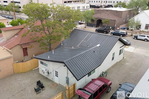 A white house with a black roof is surrounded by cars and other buildings.