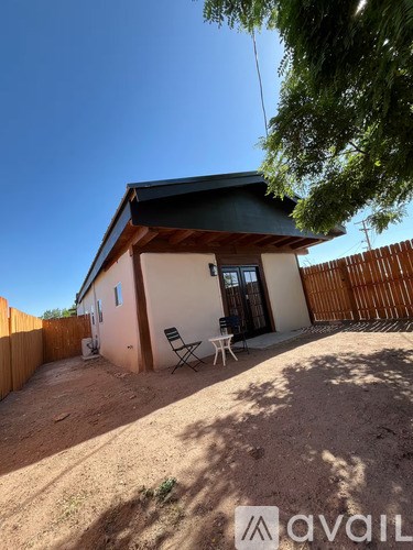 A small house with a brown roof and a white wall has a patio with a chair and table.