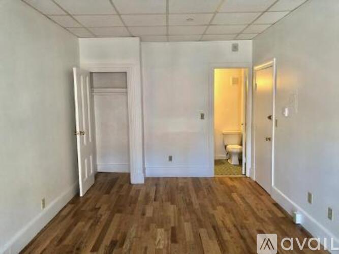 A room with wooden floors and white walls, with a toilet visible in the background.