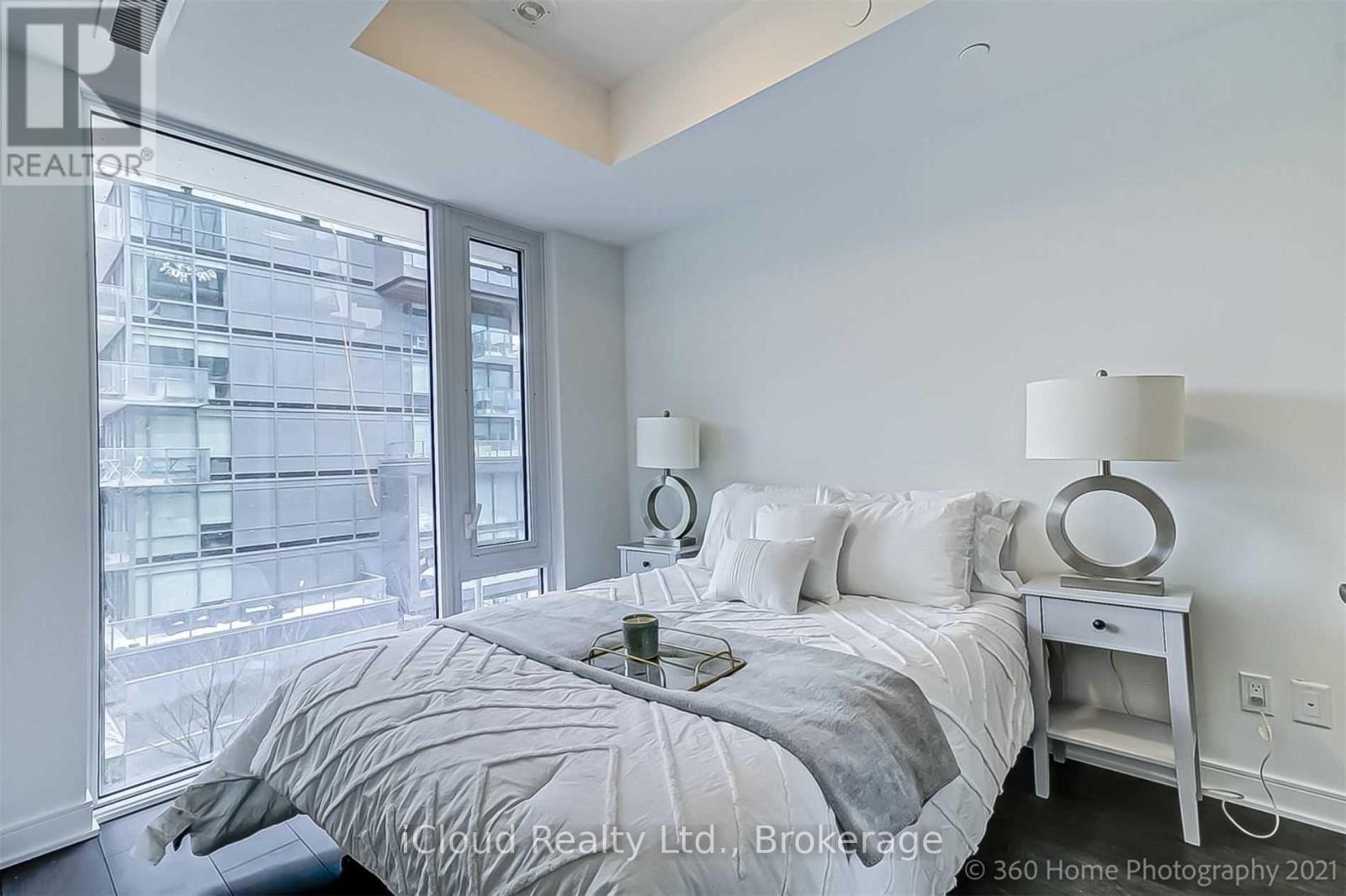 A bedroom with a large bed, a nightstand with a lamp, and a view of a cityscape through the window.