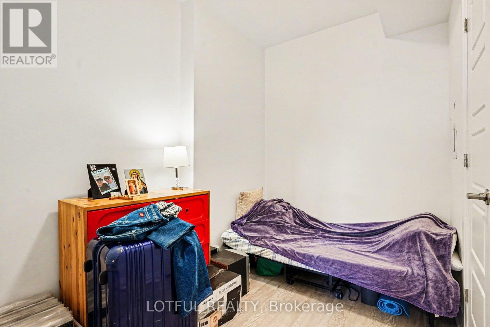 A bedroom with a bed covered in a purple blanket and a wooden cabinet with clothes on top.