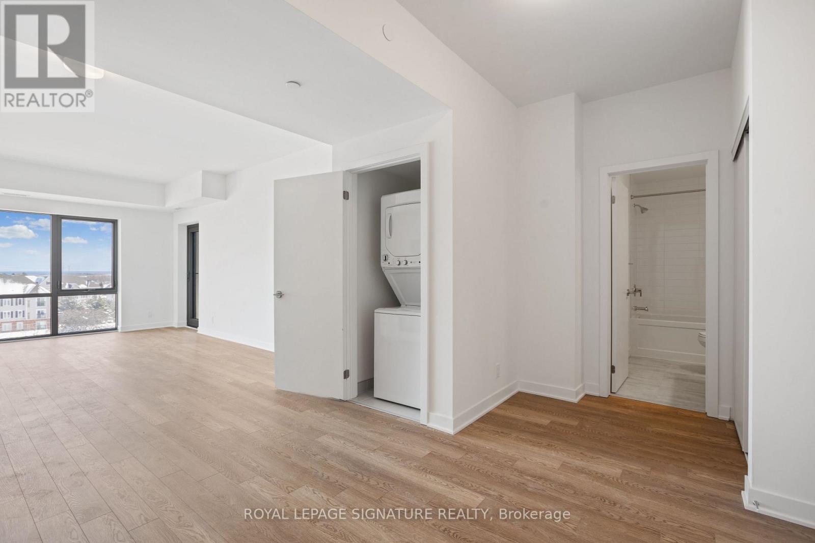 A spacious room with wooden flooring and white walls, with a large window on the left side.