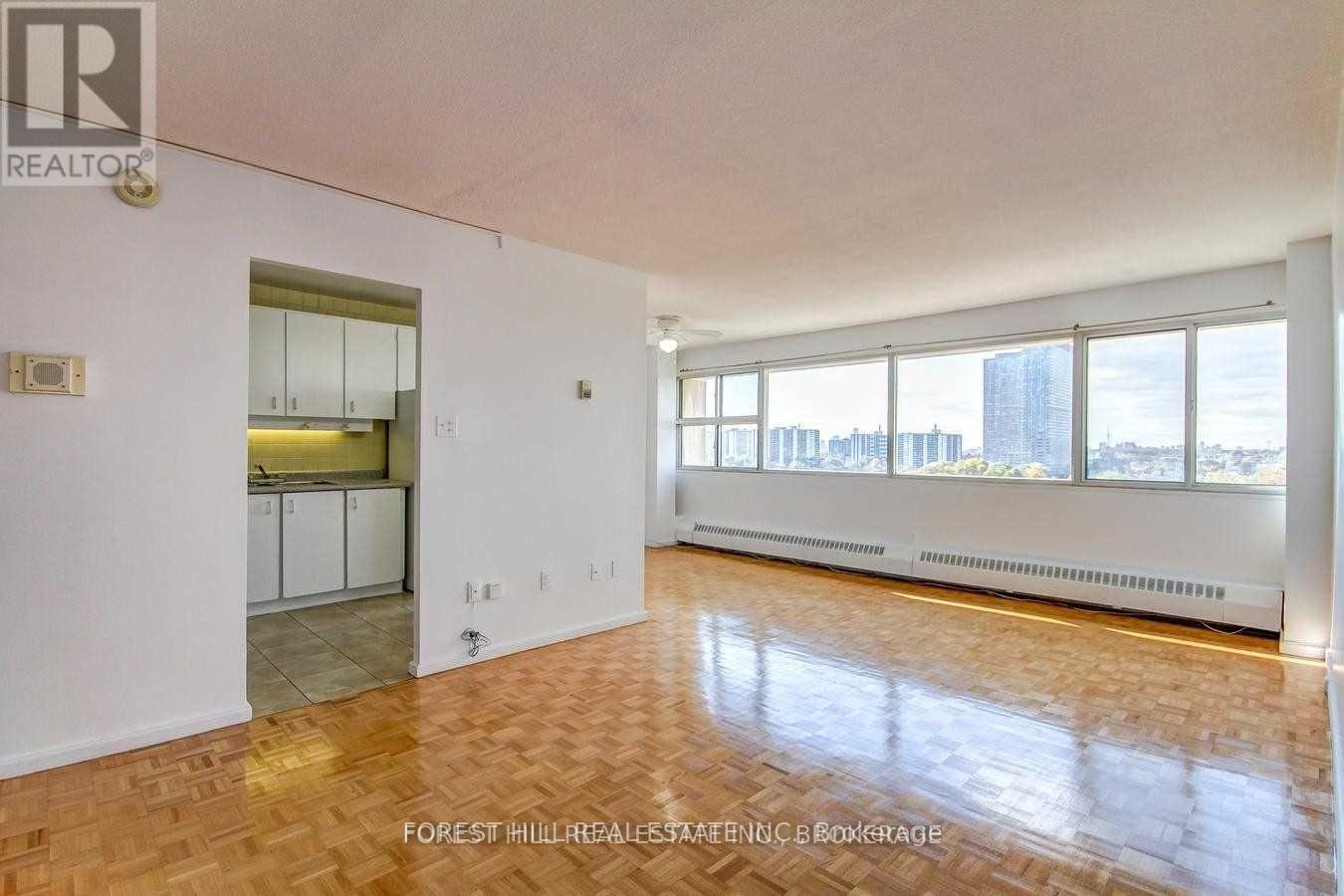 A spacious room with wooden flooring and a view of the city through large windows.