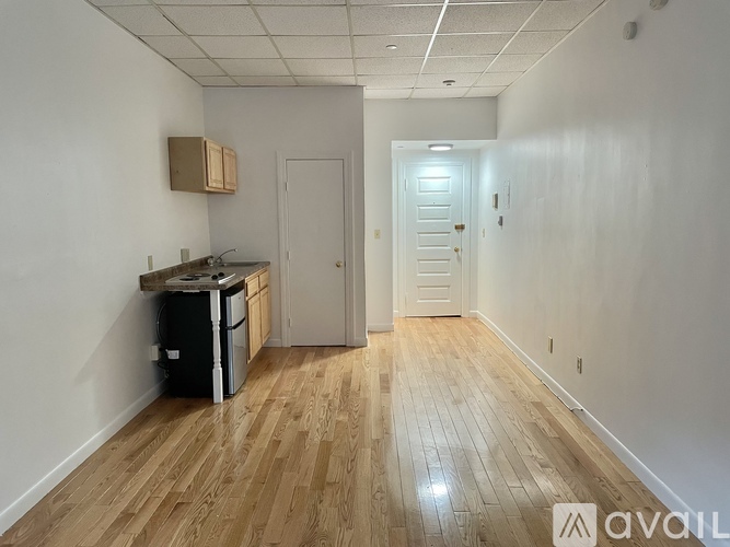 A room with wooden flooring and white walls, with a kitchenette and a door at the end.