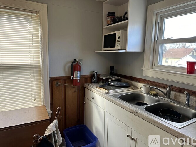 A kitchen with a sink, a window with blinds, a fire extinguisher, and a blue bin.