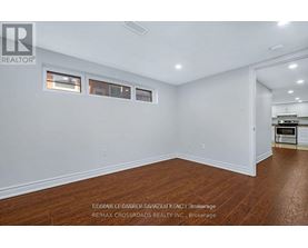 A room with white walls and wooden flooring, with a kitchen visible in the background.