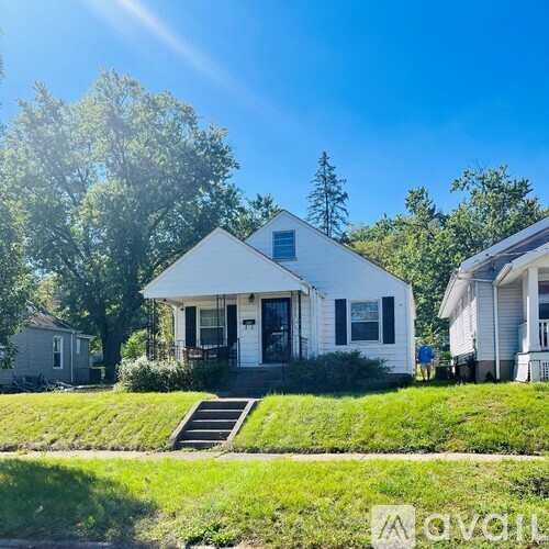 A white house with a black door and a small porch is surrounded by greenery.