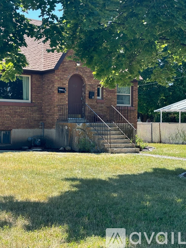A house with a brown brick exterior and a red door is surrounded by a green lawn.