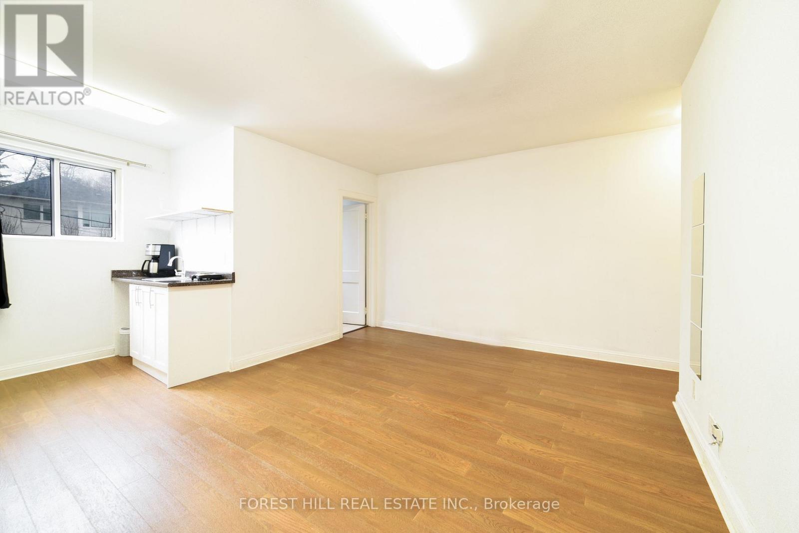 A spacious room with wooden flooring and white walls, with a window on the left side.