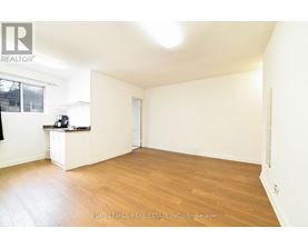 A spacious room with wooden flooring and white walls, with a window on the left side.