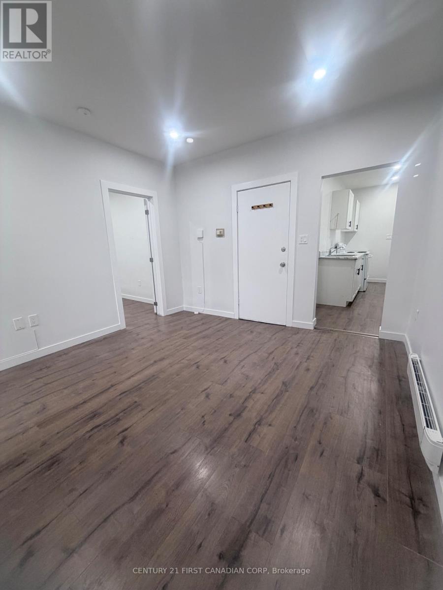 A room with wooden floors and white walls, with a door on the left and a kitchen area on the right.