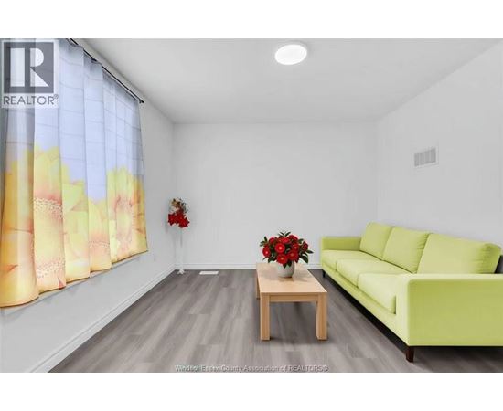 A living room with a green couch, a wooden table with a vase of red flowers, and a window with a yellow and pink curtain.