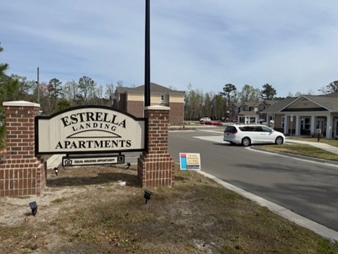 A sign for Estrella Landing Apartments stands in front of a road with a car passing by.