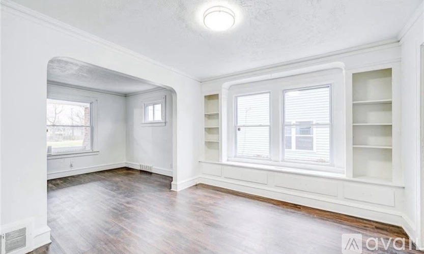 A room with white walls and wooden flooring, featuring a window with blinds and a built-in shelf.