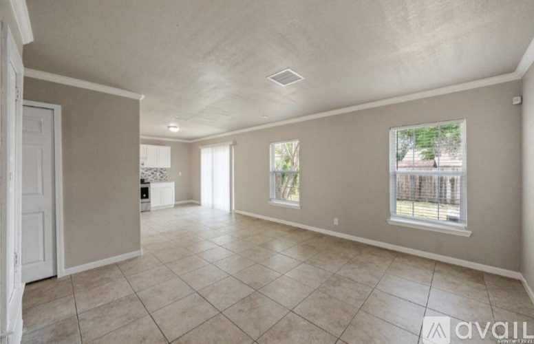 A spacious room with beige walls and tile flooring, featuring a large window with a view of a fence and greenery outside.