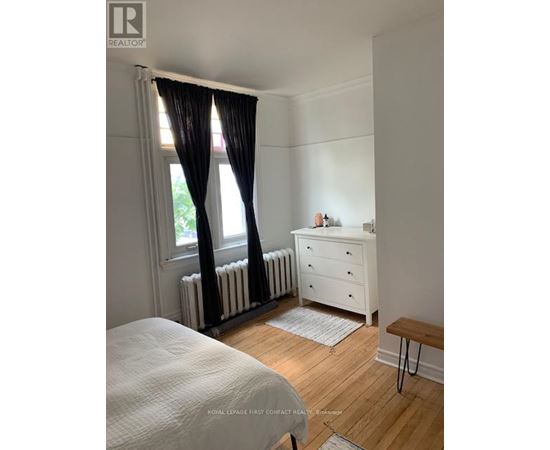 A white bedroom with a window, a radiator, a white dresser, and a wooden bench.