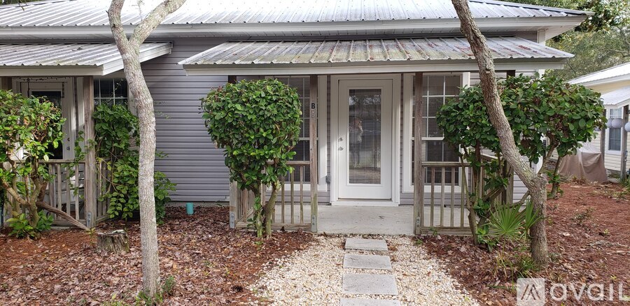 A small house with a grey roof and a white door is surrounded by trees and plants.
