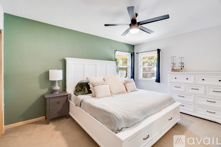 A bedroom with a white bed, a nightstand with a lamp, and a ceiling fan.