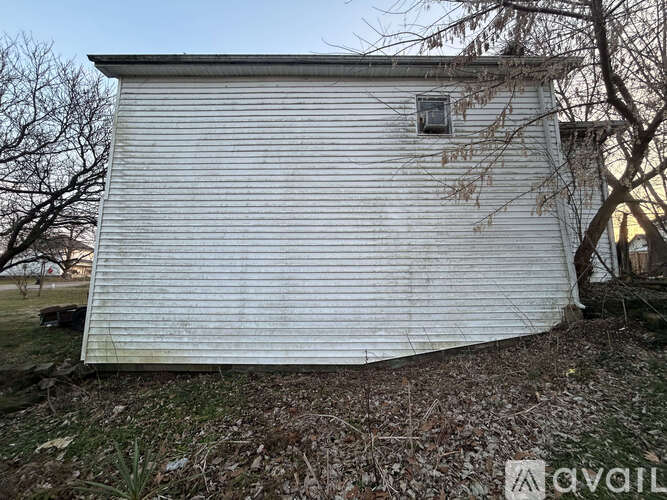 A white shed with a window and a door is surrounded by trees and grass.