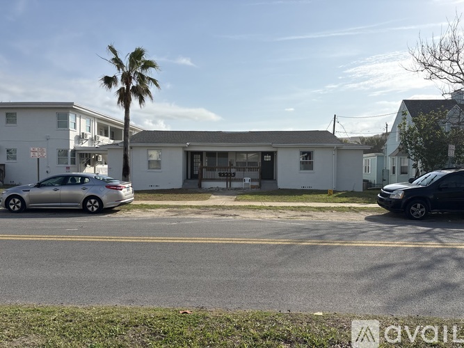 A silver car is parked on the side of the road in front of a white building with a palm tree in the background.