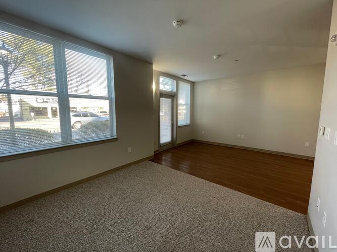A room with a carpeted floor, wooden flooring on one side, and a large window with blinds.