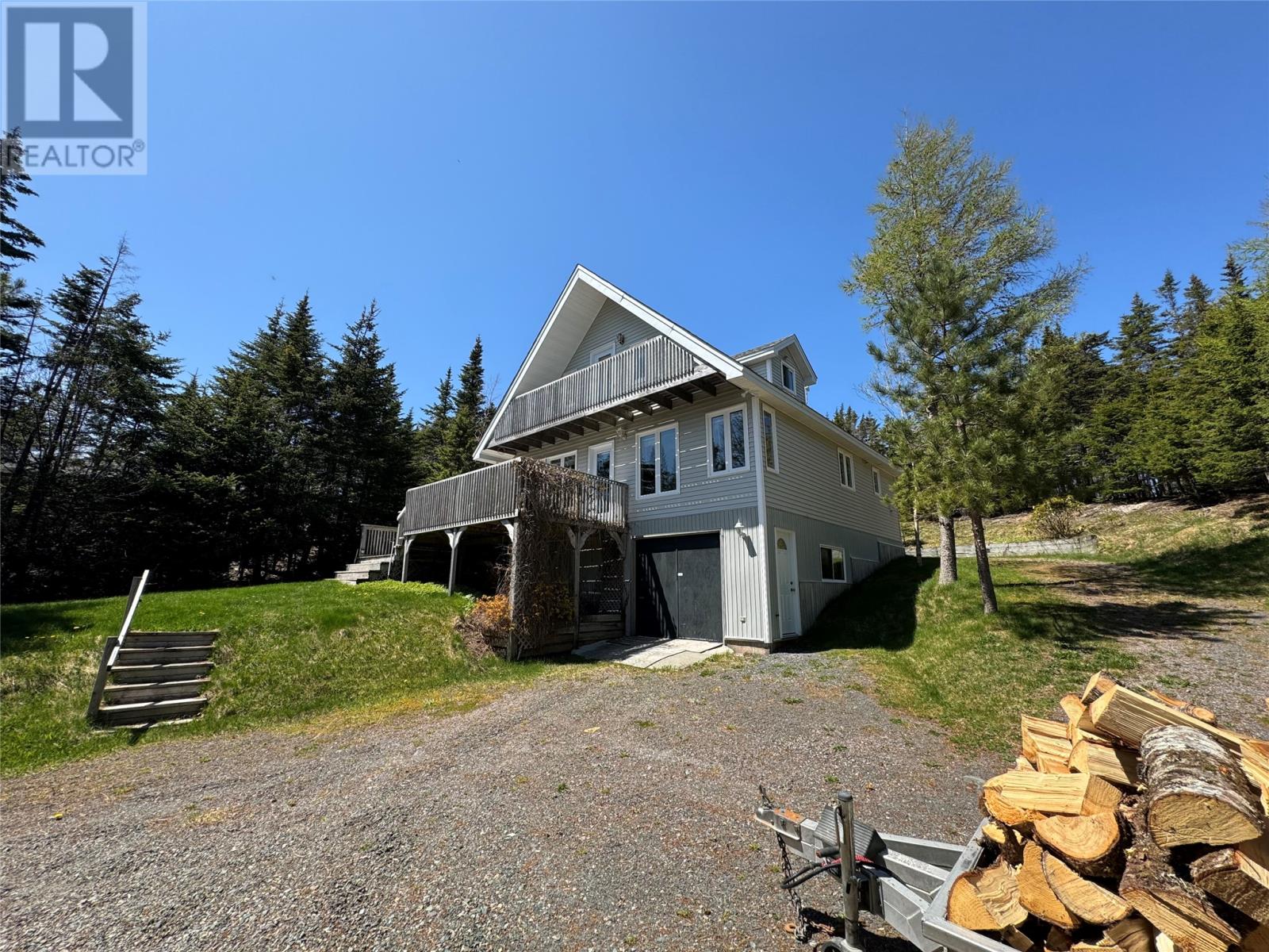 A house with a porch and a garage is surrounded by trees and has a pile of wood in front.