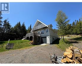 A house with a porch and a garage is surrounded by trees and has a pile of wood in front.