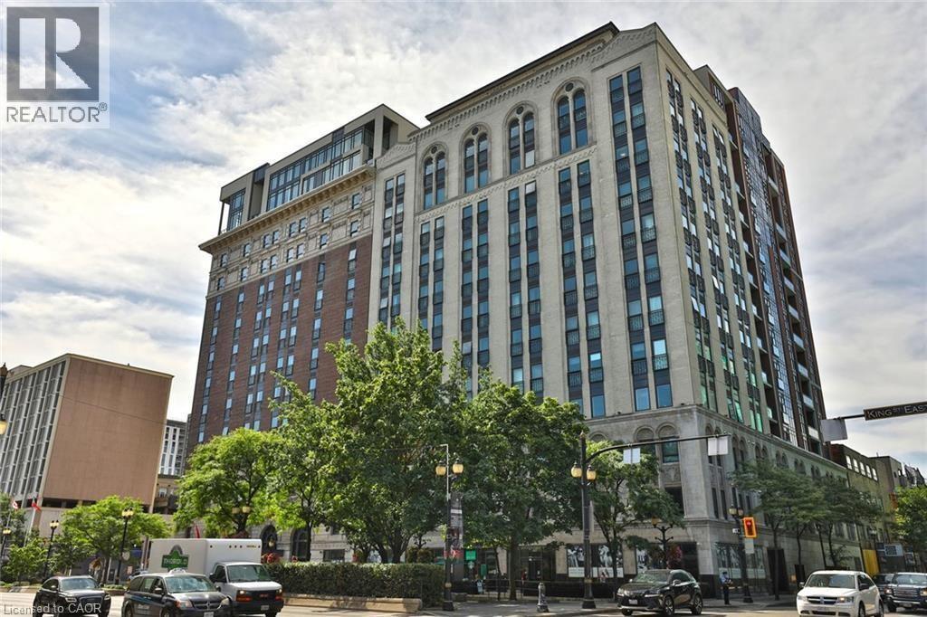A large, multi-story building with a mix of brick and glass windows is situated on a street corner with cars parked in front and a traffic light to the right.