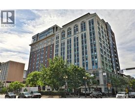 A large, multi-story building with a mix of brick and glass windows is situated on a street corner with cars parked in front and a traffic light to the right.