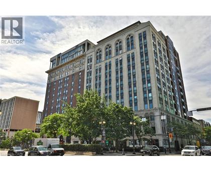 A large, multi-story building with a mix of brick and glass windows is situated on a street corner with cars parked in front and a traffic light to the right.