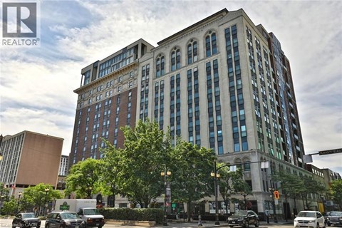 A large, multi-story building with a mix of brick and glass windows is situated on a street corner with cars parked in front and a traffic light to the right.