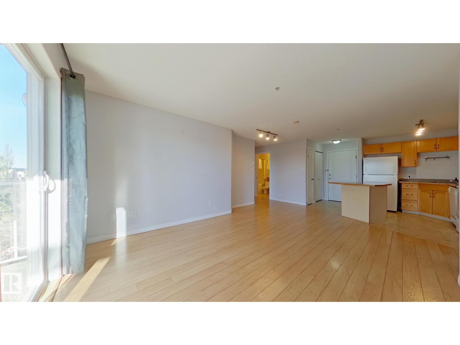 A room with wooden floors and a kitchen area in the background.