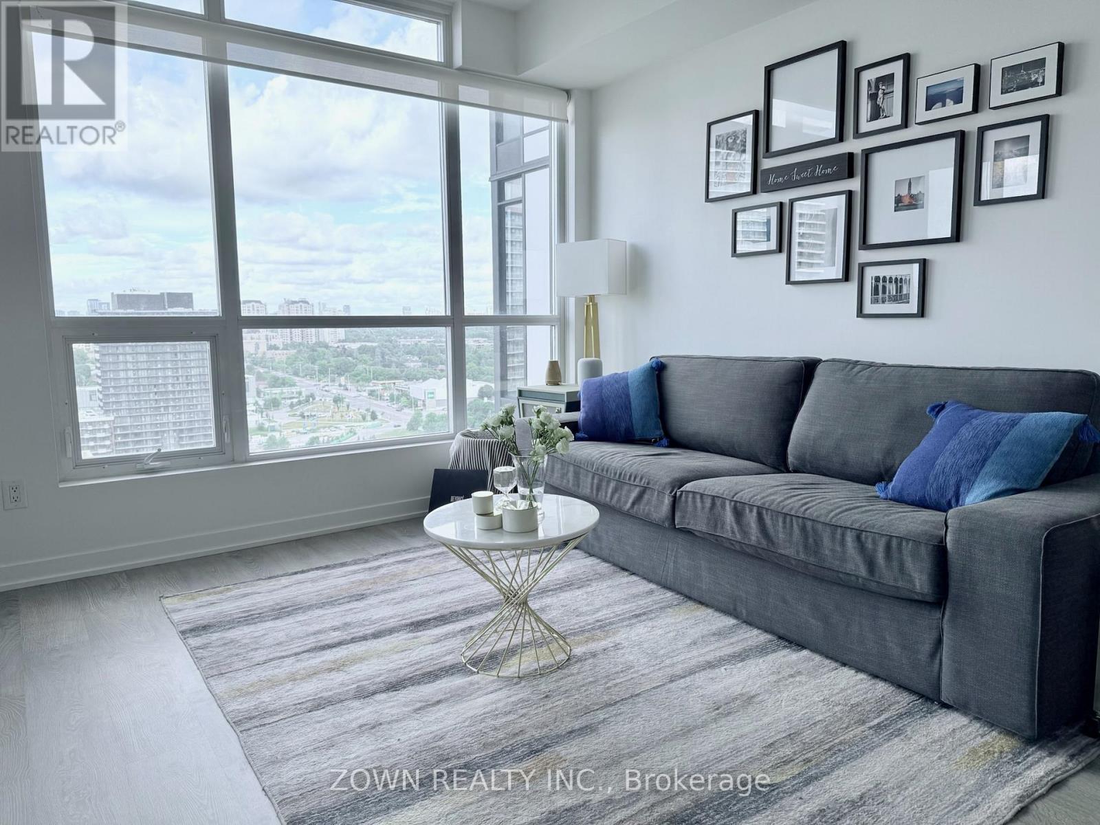 A living room with a grey couch, a round table, and a view of the city through the windows.
