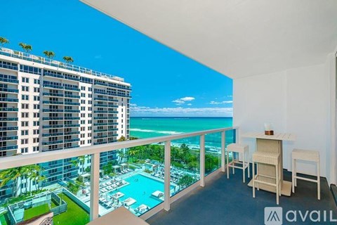 A balcony with chairs and a table overlooks a pool and a beachfront building.